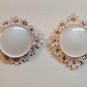 May include: A pair of vintage clip-on earrings. Each earring features a large, round, white center stone surrounded by small, light pink flower-shaped accents. Clear rhinestones are interspersed among the flowers, adding sparkle. The earrings have a diamond-like shape.