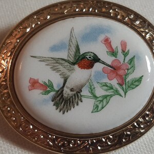 May include: An oval brooch featuring a gold-toned metal frame with an intricate embossed border. The centre is a white porcelain-like inlay depicting a hummingbird with green, white, and red plumage hovering near pink flowers against a blue sky.