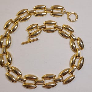 May include: A gold-toned necklace featuring a series of polished, elongated oval links connected by smaller rectangular bars. The necklace is secured with a toggle clasp consisting of a circular ring and a straight bar, creating a continuous chain.