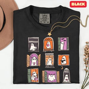 May include: Black t-shirt with a whimsical design of ghosts in windows. Each window features a different ghost, some reading, listening to music, or with a cat. The shirt has a Comfort Colors tag and a gold star necklace. The text "The Cozy Haus" is also on the shirt.