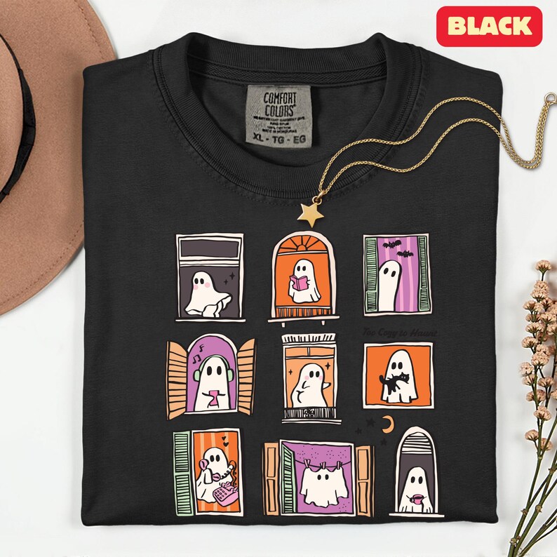 May include: Black t-shirt with a whimsical design of ghosts in windows. Each window features a different ghost, some reading, listening to music, or with a cat. The shirt has a Comfort Colors tag and a gold star necklace. The text "The Cozy Haus" is also on the shirt.