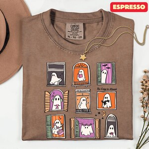 May include: Espresso-colored Comfort Colors t-shirt with a whimsical ghost window design. The shirt features nine illustrated windows, each with a unique ghost scene. The text "Too Cozy to Haunt" is printed on the shirt. A gold star pendant necklace is draped on the shirt.