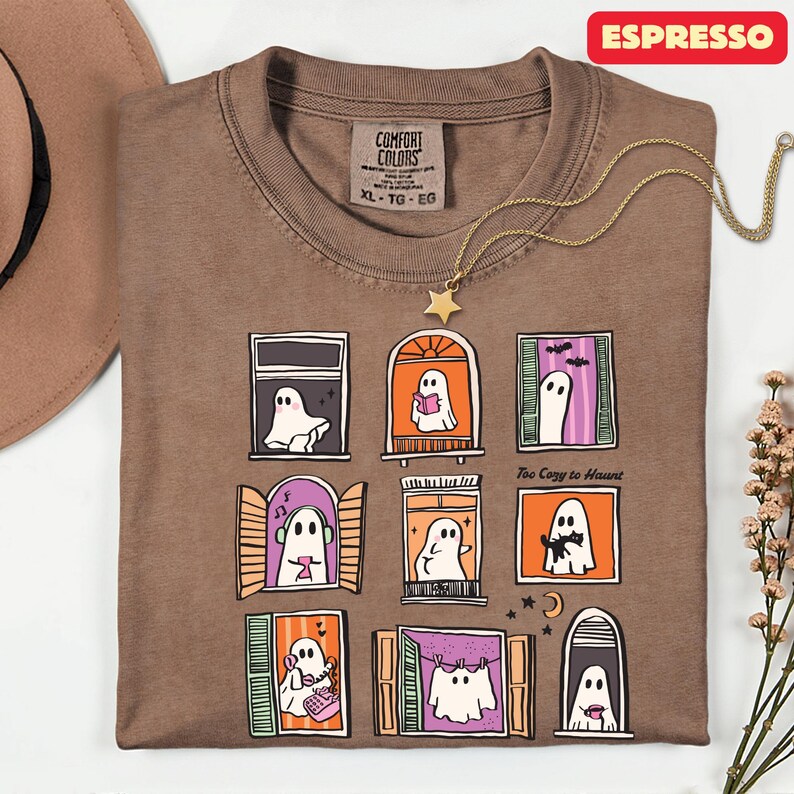 May include: Espresso-colored Comfort Colors t-shirt with a whimsical ghost window design. The shirt features nine illustrated windows, each with a unique ghost scene. The text "Too Cozy to Haunt" is printed on the shirt. A gold star pendant necklace is draped on the shirt.