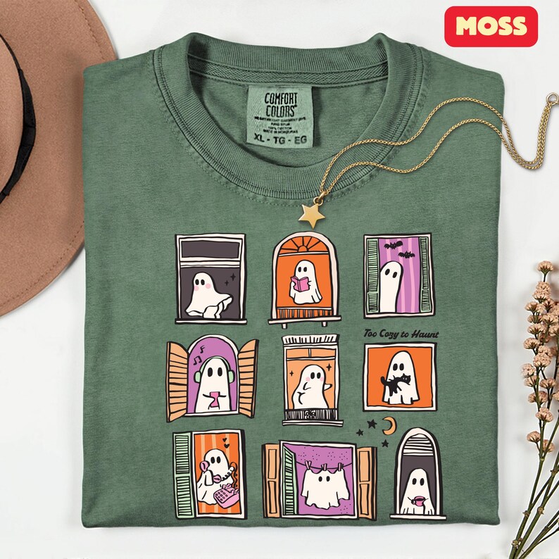 May include: Green XL Comfort Colors t-shirt with a graphic of cartoon ghosts in windows. The shirt has the text "Too Cozy to Haunt". A gold necklace with a star pendant is draped across the shirt.