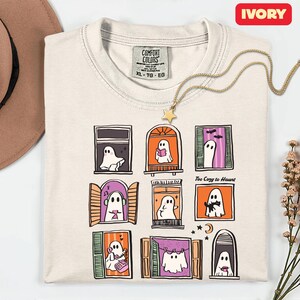 May include: Ivory-colored t-shirt with a whimsical ghost window design. The shirt features a grid of nine windows, each showcasing a different ghost scene. The text "Too Cozy to Haunt" is printed on the shirt.