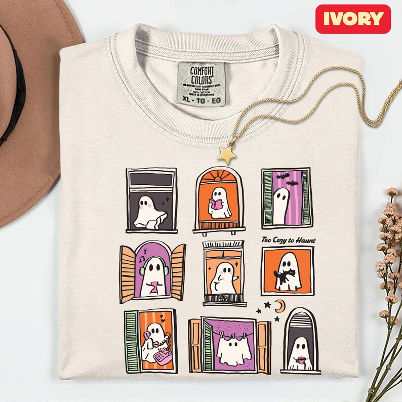 May include: Ivory-colored t-shirt with a whimsical ghost window design. The shirt features a grid of nine windows, each showcasing a different ghost scene. The text "Too Cozy to Haunt" is printed on the shirt.