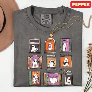 May include: Dark gray Comfort Colors t-shirt with a whimsical Halloween ghost window design. The shirt features nine illustrated windows with ghosts, stars, and the text "Too Cozy to Haunt". A gold star pendant necklace is draped across the shirt.