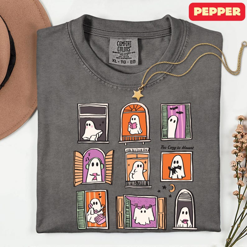 May include: Dark gray Comfort Colors t-shirt with a whimsical Halloween ghost window design. The shirt features nine illustrated windows with ghosts, stars, and the text "Too Cozy to Haunt". A gold star pendant necklace is draped across the shirt.