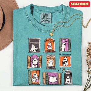 May include: Seafoam green t-shirt with a whimsical ghost window design. Each window features a different ghost illustration. A gold star pendant necklace rests on the shirt. The text "Too Cozy to Haunt" is printed on the shirt.