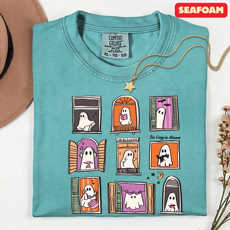 May include: Seafoam green t-shirt with a whimsical ghost window design. Each window features a different ghost illustration. A gold star pendant necklace rests on the shirt. The text "Too Cozy to Haunt" is printed on the shirt.