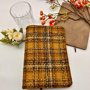 Puede incluir: Una cubierta de diario a cuadros dorados y negros con un marcador de cinta marrón. También hay un vaso transparente estampado y un cuaderno marrón con un marcador rojo. Flores decorativas y ramas con bayas naranjas completan la escena.