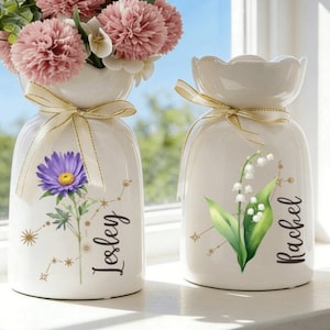 Puede incluir: Dos jarrones de cerámica blanca con diseños florales y lazos dorados. Uno presenta una flor morada y el nombre "Lesley", el otro un lirio de los valles y el nombre "Rachel". Ambos jarrones contienen flores rosas.