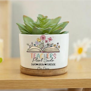 Puede incluir: Una maceta de cerámica blanca con el texto "TEACHERS Plant Seeds THAT GROW FOREVER" y un diseño floral. La maceta contiene una planta suculenta verde y se apoya en un posavasos redondo de madera. El texto "Mrs. Davis" está debajo.