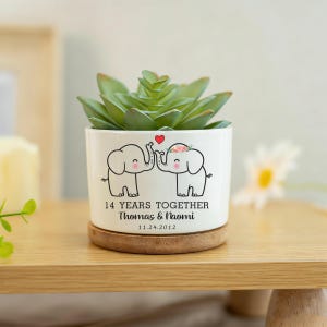 May include: A white ceramic planter with a succulent plant. The planter features two cartoon elephants touching trunks under a red heart. Text reads "14 YEARS TOGETHER" with names and a date. The planter sits on a wooden coaster.