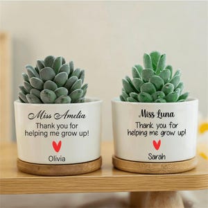 Puede incluir: Dos macetas de cerámica blanca con suculentas, cada una sobre una base de madera. Las macetas tienen el texto "Miss Amelia" y "Miss Luna" con el mensaje "¡Gracias por ayudarme a crecer!" y un corazón rojo. Los nombres "Olivia" y "Sarah" están debajo.