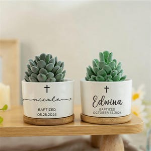 May include: Two white ceramic planters with succulents, each on a wooden coaster. One planter has the name "nicole" and the date "05.25.2025", the other has "Edwina" and "OCTOBER 12.2024". Both have a cross symbol.
