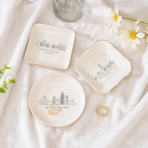 May include: Three white ceramic ring dishes with gold rims. Each dish features a black skyline design and personalized text. One square dish reads "NANCY & LEO 09.20.2025". A round dish says "ALBERTA & MAX 11.06.2025". A fourth dish is also included.