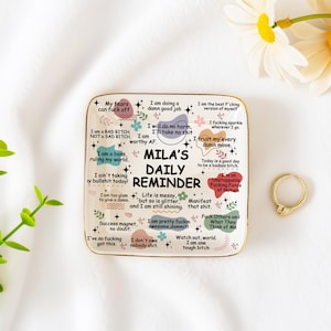 Könnte beinhalten: Ein quadratisches Keramik-Schmucktablett mit goldenem Rand, mit dem Text "MILA'S DAILY REMINDER" und verschiedenen inspirierenden Sätzen. Rechts befindet sich ein goldener Ring mit einem kleinen rechteckigen Stein. Das Tablett steht auf einer weißen Oberfläche.