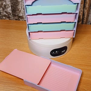 Puede incluir: Una caja de almacenamiento de plástico de color arcoíris pastel con cuatro cajones y una bandeja extraíble. La caja tiene una base blanca con el texto "CWOCH/COW" y "DC-5V".