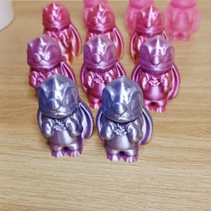 Könnte beinhalten: Eine Ansammlung kleiner, metallisch aussehender Figuren in Rosa- und Lilatönen. Die Figuren haben ein cartoonartiges Design mit großen Ohren und einem runden Körper. Sie sind auf einer hellen Holzoberfläche angeordnet.