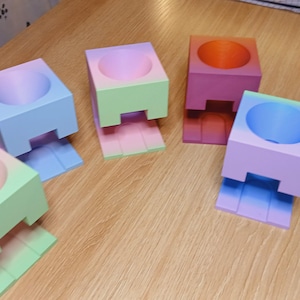 May include: Five ombre planter stands in different colours. The stands are made of a smooth, solid material and have a square base with a round top. The colours are light blue, light green, pink, red, and purple.