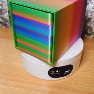 Puede incluir: Una caja multicapa de colores arcoíris con una capa exterior verde. La caja está hecha de plástico y tiene un acabado liso y brillante.