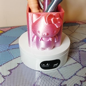 May include: A pink 3D-printed organizer shaped like a cartoon cat, holding various craft tools. The organizer sits on a white rotating display stand. Tools include pens and styluses.
