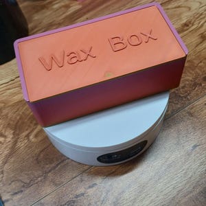 Puede incluir: Caja de cera rectangular naranja con las palabras "Wax Box" en relieve en la parte superior, con un borde rosa. Descansa sobre una base circular blanca. El fondo muestra una superficie de madera y un objeto rosa.