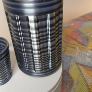 May include: A cylindrical, textured container with a dark gray and silver striped pattern. The container has a ridged top and is set on a white surface. A smaller version of the container is visible to the left.