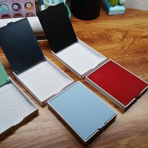 May include: An assortment of open, hinged plastic trays in various colors, including green, black, red, and blue. Each tray has a white interior with a ridged design. The trays are arranged on a wooden surface.