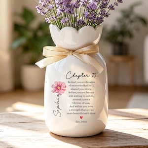 May include: A white ceramic vase with a scalloped rim and a beige ribbon tied around the neck, filled with purple lavender sprigs. The vase features the text "Chapter 70", a pink flower illustration, the name "Sophia", and the date "Est. 1956".