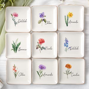 Könnte beinhalten: Neun quadratische, cremefarbene Keramikschalen mit goldenen Rändern, jede mit einem anderen Aquarell-Blumendesign und einem Namen in schwarzer Schrift. Zu den Blumen gehören Nelken, Veilchen, Narzissen, Rosen und mehr. Namen sind Delilah, Chloe, Amanda, Bonnie, Nicole und Cecilia.