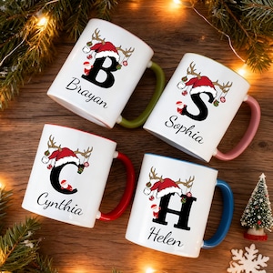 以下が含まれることがあります： パーソナライズされたクリスマスデザインの白いセラミックマグ4個。各マグには、文字、名前、サンタ帽とトナカイの角のイラストが描かれています。マグには、緑、ピンク、赤、青など、さまざまな色のハンドルと内側の縁があります。