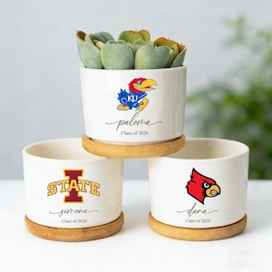 May include: Three white ceramic planters with wooden saucers, each featuring a college logo and a name with "Class of 2026". One planter holds a succulent. The logos include a red cardinal, a blue and yellow Jayhawk, and a gold and maroon "STATE" logo.