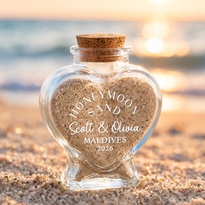 May include: A heart-shaped clear glass bottle with a cork stopper, filled with light tan sand. The front of the glass features white text that reads HONEYMOON SAND, Scott & Olivia, MALDIVES, and 2026, printed in a mix of serif and script fonts.