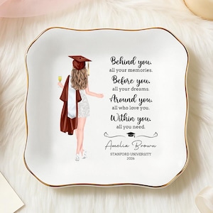 May include: A white ceramic trinket dish with a gold rim, featuring a graduation-themed illustration and inspirational text. The dish is surrounded by decorative elements like roses, a candle, and jewelry.