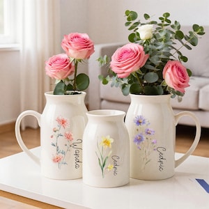 Puede incluir: Tres jarrones de cerámica blanquecina con diseños florales y los nombres "Wanda" y "Cedric" impresos. Los jarrones contienen rosas rosas y vegetación. Un jarrón tiene un asa.