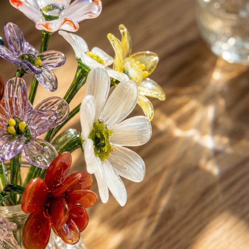 May include: A bouquet of colorful glass flowers in a clear vase. The flowers include white, yellow, purple, and red blooms with green stems. The glass is translucent, allowing light to pass through, creating a delicate and artistic display.