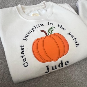 May include: Cream-coloured sweatshirt featuring an embroidered orange pumpkin design. The text "Cutest pumpkin in the patch" arches around the pumpkin, with the name "Jude" below. A seasonal autumn-themed garment.