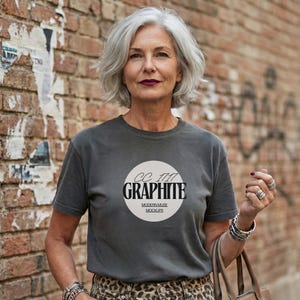 May include: A woman wearing a gray t-shirt with a circular graphic that reads "CC 1111 GRAPHITE MODERN MUSE MOCKUPS". She is also wearing leopard print pants, silver rings, bracelets, and carrying a brown tote bag. The background is a brick wall.