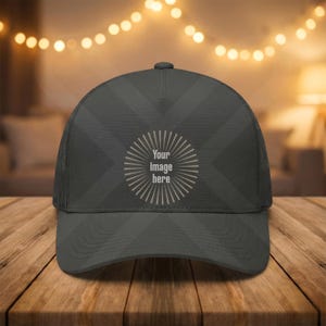 May include: A dark gray trucker hat with a mesh back and a curved brim. The front of the hat features a circular design with the text "Your image here" in a light gray color. The hat is displayed on a wooden surface.