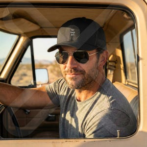 May include: A black trucker hat with a mesh back and a curved brim. The hat features a circular design with the text "your image here". The person in the image is wearing aviator sunglasses and a gray t-shirt.