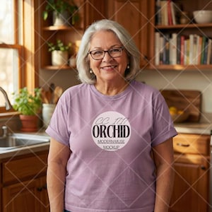 May include: A woman wearing a lilac-colored t-shirt with rolled-up sleeves. The shirt features a circular graphic with the text "CC 1717 ORCHID MODERN MUSE MOCKUP". She is wearing glasses and smiling. The background includes a kitchen with wooden cabinets and a window.