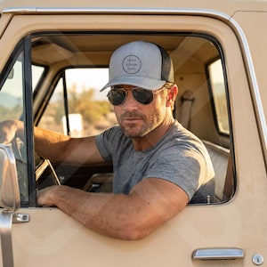 May include: A gray and black trucker hat with the text "your image here" on the front. The hat is worn by a person wearing sunglasses and a gray t-shirt, sitting in a tan vehicle.