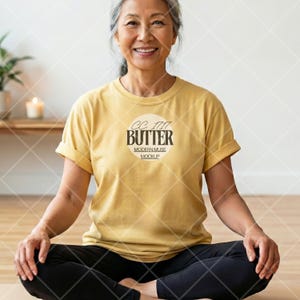 Può includere: Una donna indossa una maglietta giallo senape con le maniche arrotolate e la scritta "BUTTER" stampata sul davanti. La donna è seduta a gambe incrociate, indossa pantaloni neri. La maglietta ha uno scollo tondo.