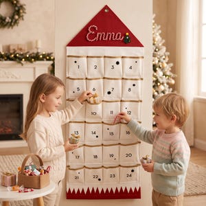 Pode incluir: Um calendário do advento de tecido para pendurar na parede com vinte e quatro bolsos brancos, numerados de um a vinte e quatro. O topo apresenta um telhado triangular vermelho com o nome Emma em branco e um pequeno enfeite de árvore.