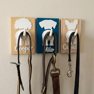 May include: Three rectangular wall-mounted leash holders featuring white 3D dog rear silhouettes with protruding tails as hooks. The left plaque is natural wood with the name Cooper, the center is blue with Milo, and the right is natural wood with Cookie.