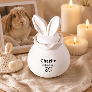 Puede incluir: Una urna de cerámica blanca con forma de conejito, personalizada con el nombre "Charlie", las fechas 2018-2025 y un corazón con huella de pata. Una foto enmarcada de un conejo, un gorro de conejito de ganchillo y velas encendidas en el fondo.