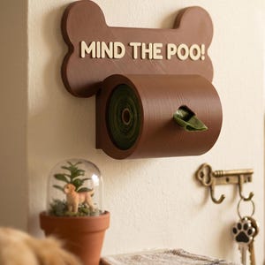 May include: A brown wall-mounted dog waste bag dispenser shaped like a bone with the text MIND THE POO! in cream-colored letters. The dispenser holds a roll of dark green waste bags, with one bag partially pulled through the center opening.