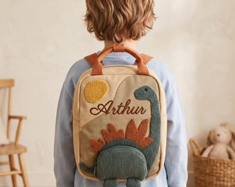 May include: A beige backpack featuring a textured green dinosaur appliqué with orange spikes, a yellow sun, and the embroidered name Arthur. The bag includes brown top handles and shoulder straps, with a zippered front pocket compartment.
